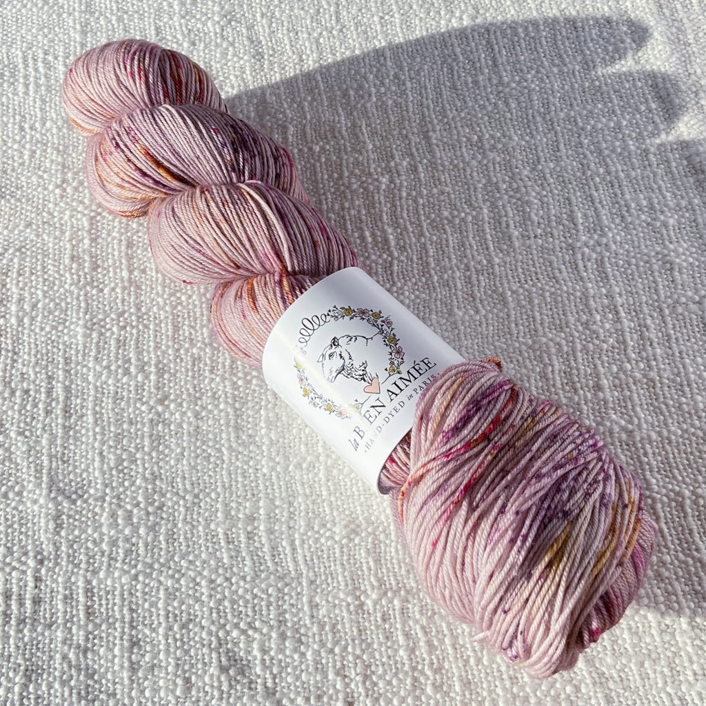 An image of a skein of sock yarn in a pale pink base with speckles of pink, purple, and yellow, with a white ‘La Bien Aimee’ label against a canvas background.