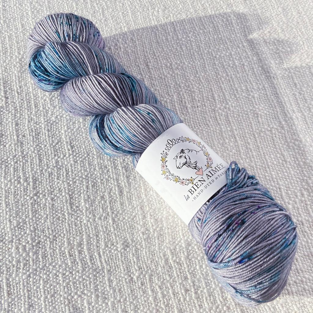 An image of a skein of sock yarn in a pale purple gray base with speckles of teal, navy, and purple, with a white ‘La Bien Aimee’ label against a canvas background.