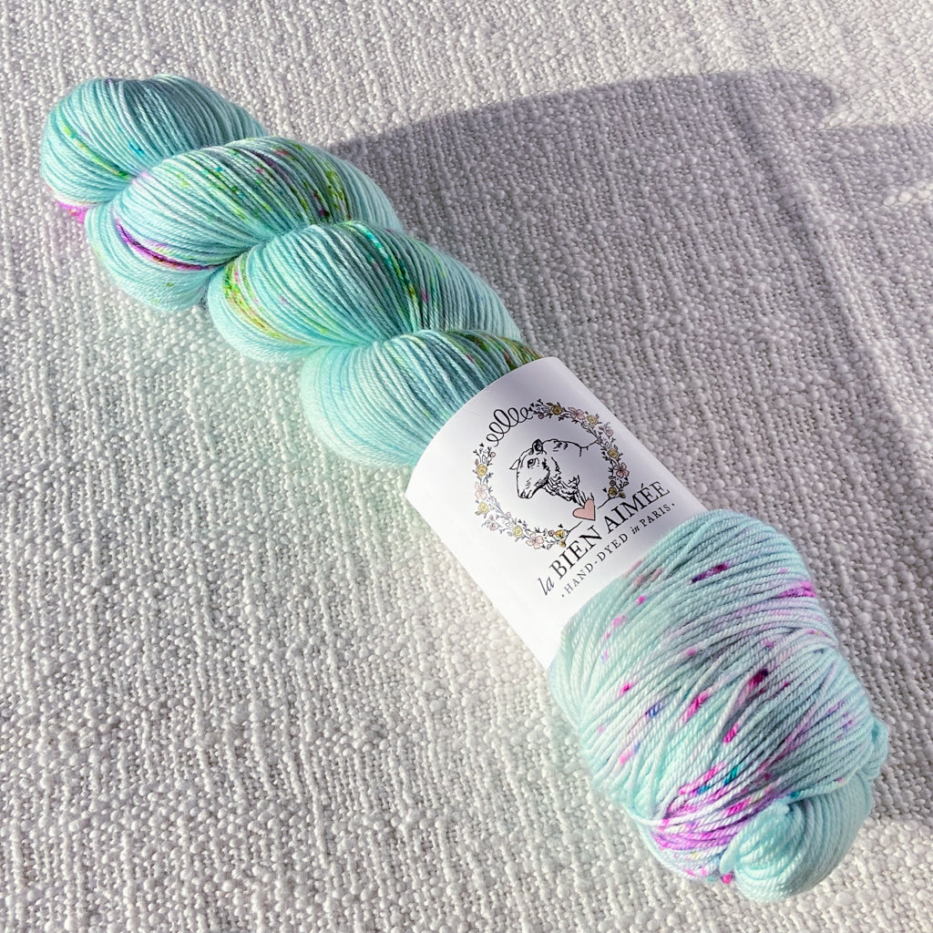 An image of a skein of sock yarn in an aqua blue base with speckles of pink and green, with a white ‘La Bien Aimee’ label against a canvas background.