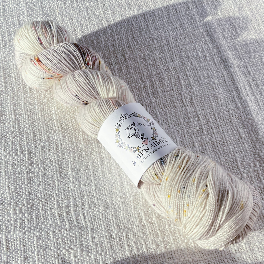 An image of a skein of sock yarn in an off-white color with speckles of orange, teal, and yellow, with a white ‘La Bien Aimee’ label against a canvas background.