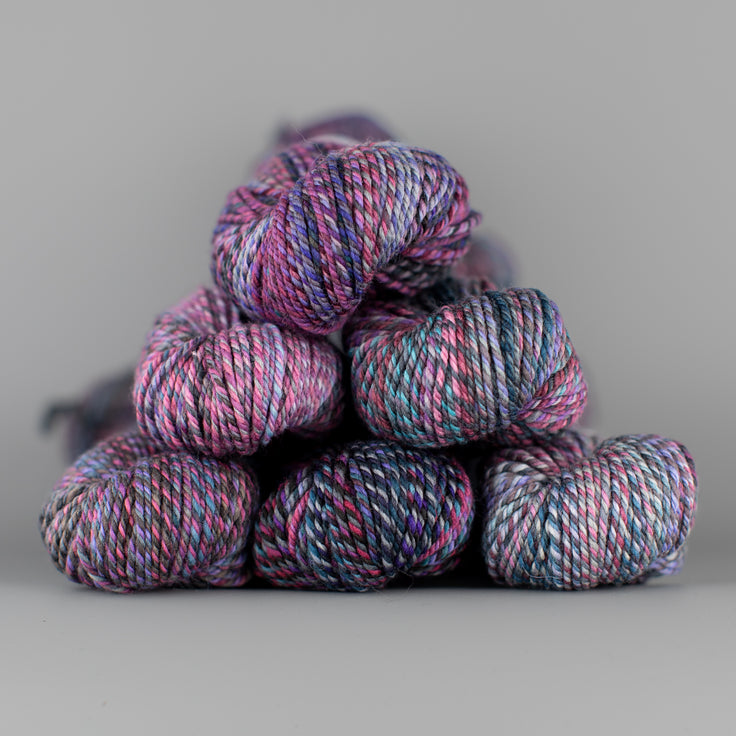 An image of skeins of Spincycle Dream State worsted weight yarn in a soft pink, purple, blue, and charcoal color gradient against a gray background.