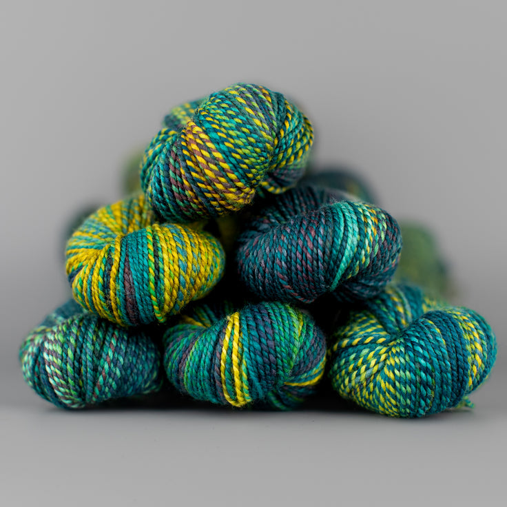 An image of skeins of Spincycle Dream State worsted weight yarn in a teal, yellow, and green color gradient against a gray background.
