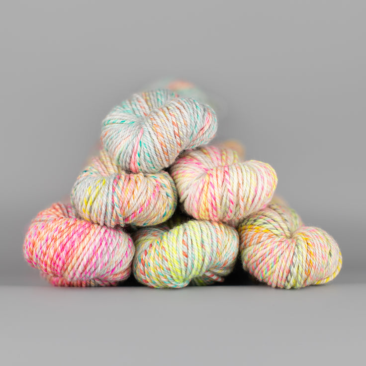 An image of skeins of Spincycle Dream State worsted weight yarn in a pale orange, yellow, pink, and blue gray color gradient against a gray background.