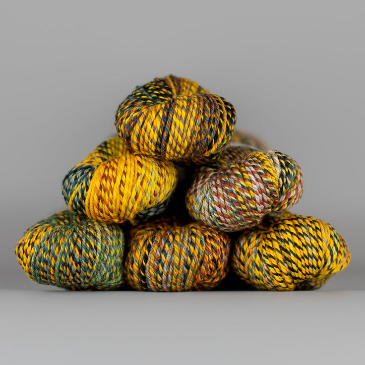 An image of skeins of Spincycle Dream State worsted weight yarn in a yellow, green, copper, and charcoal color gradient against a gray background.