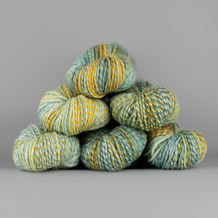 An image of skeins of Spincycle Dream State worsted weight yarn in a yellow and blue green color gradient against a gray background.