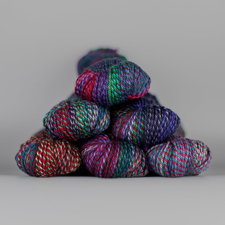 An image of skeins of Spincycle Dream State worsted weight yarn in a red, purple, green, and charcoal color gradient against a gray background.