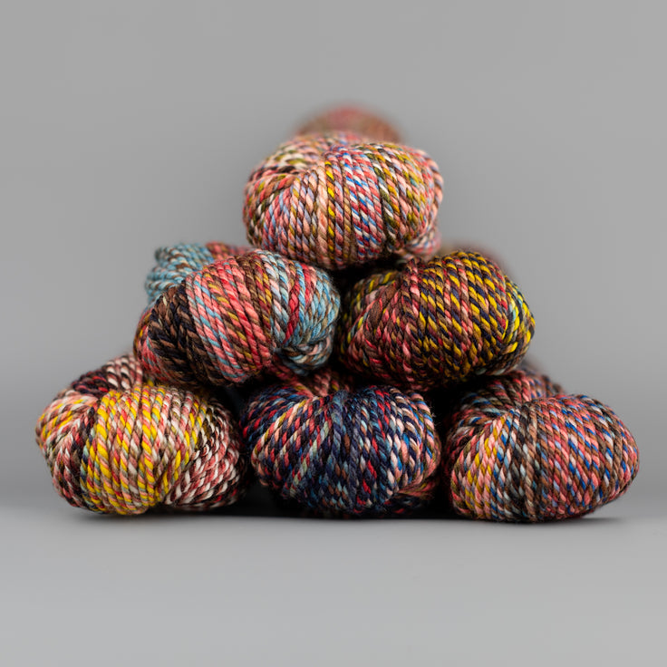 An image of skeins of Spincycle Dream State worsted weight yarn in a pink, yellow orange, blue and brown color gradient against a gray background.