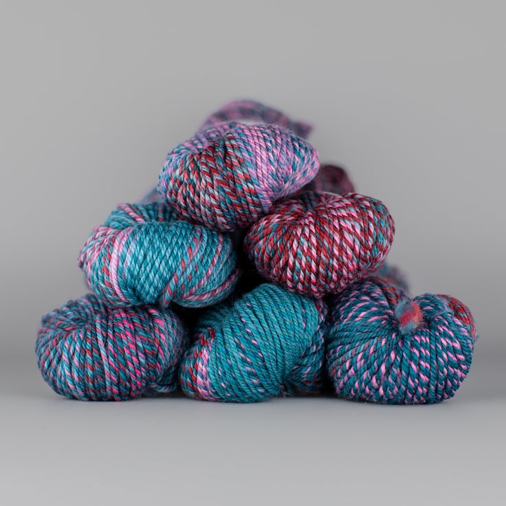 An image of skeins of Spincycle Dream State worsted weight yarn in a teal, pink, and red color gradient against a gray background.