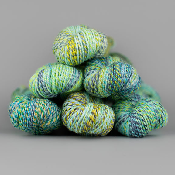 An image of skeins of Spincycle Dream State worsted weight yarn in a pale green, blue, and gray color gradient against a gray background.