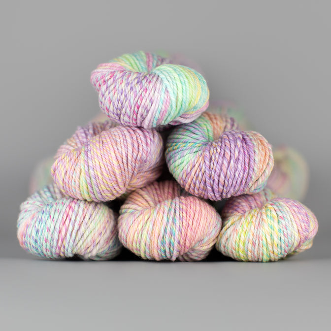 An image of skeins of Spincycle Dream State worsted weight yarn in a pastel blue, peach, purple, and yellow color gradient against a gray background.