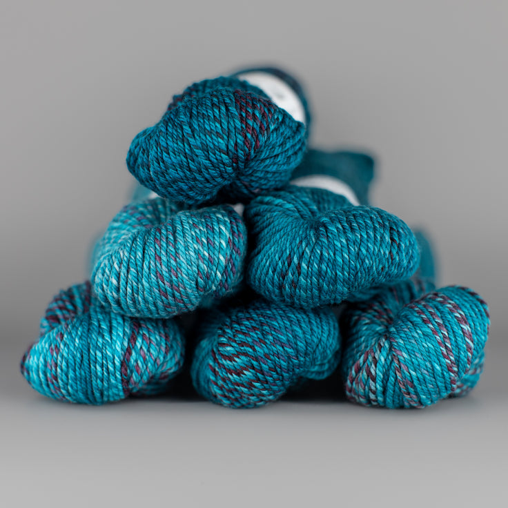 An image of skeins of Spincycle Dream State worsted weight yarn in a teal and deep purple color gradient against a gray background.