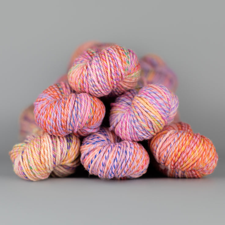 An image of skeins of Spincycle Dream State worsted weight yarn in an orange, pink, and purple color gradient against a gray background.