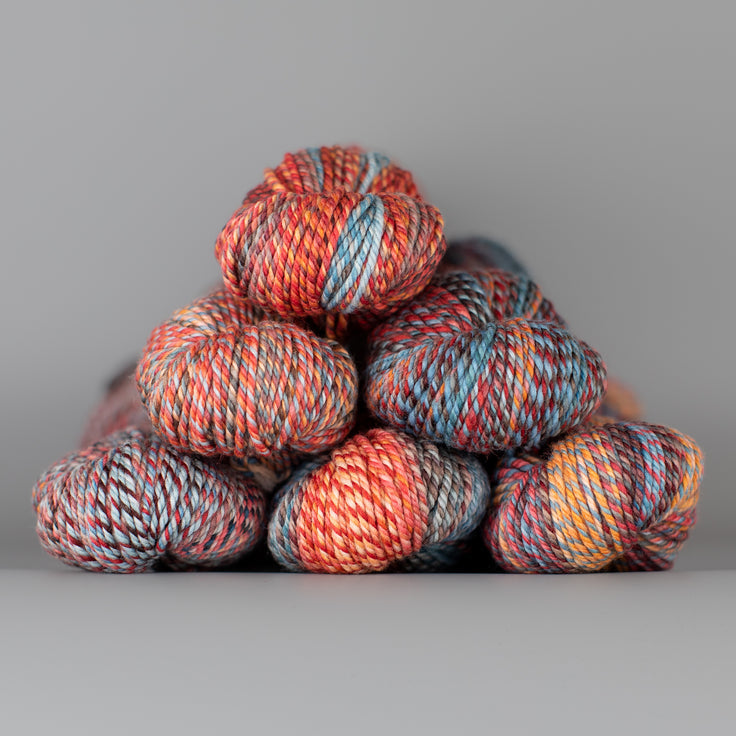 An image of skeins of Spincycle Dream State worsted weight yarn in a red orange, blue, and brown color gradient against a gray background.