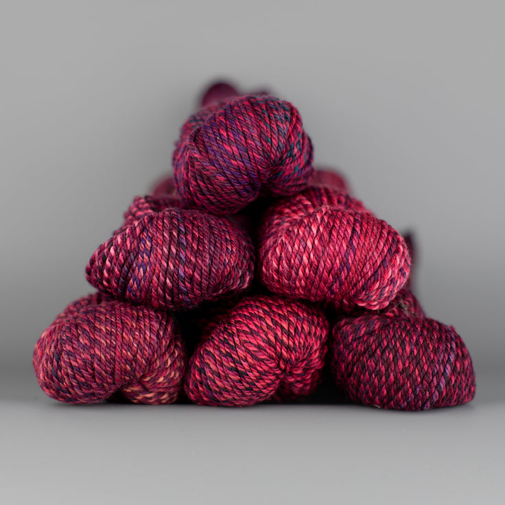 An image of skeins of Spincycle Dream State worsted weight yarn in a magenta, purple, and red color gradient against a gray background.