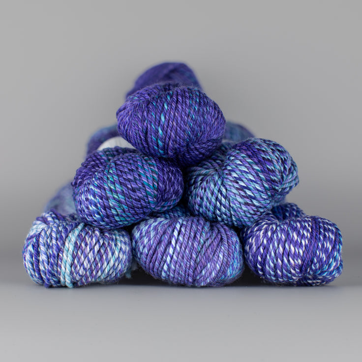 An image of skeins of Spincycle Dream State worsted weight yarn in a dark purple and bright blue color gradient against a gray background.
