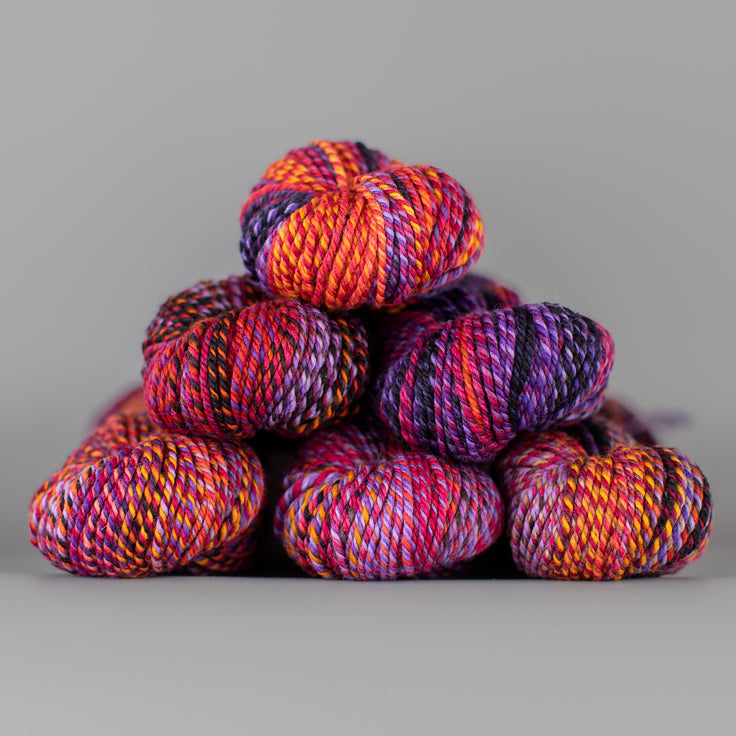 An image of skeins of Spincycle Dream State worsted weight yarn in an orange, pink, and purple color gradient against a gray background.