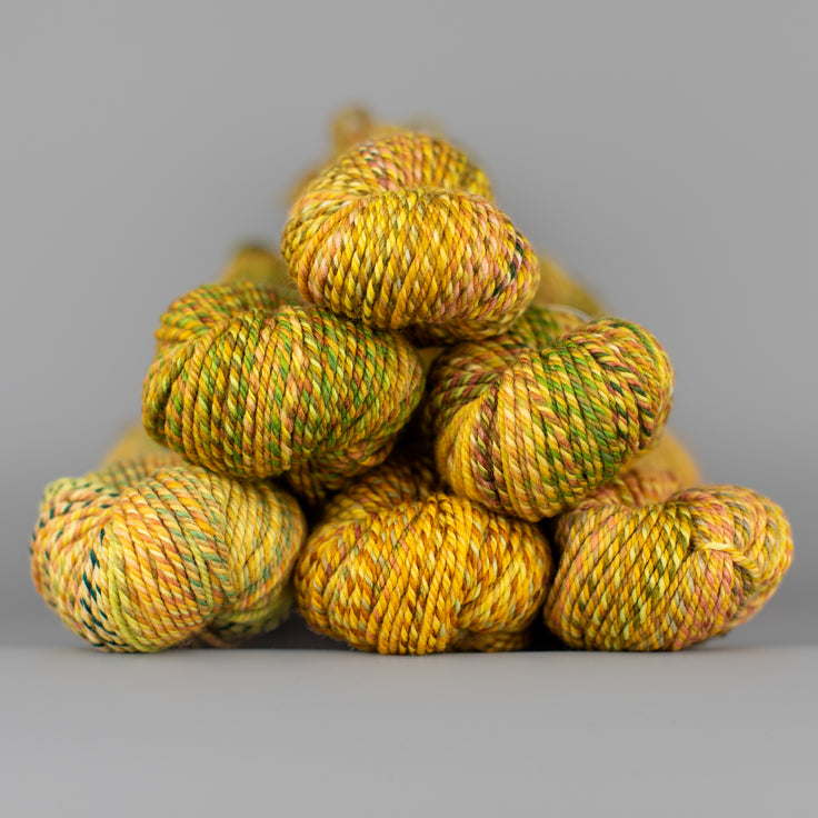 An image of skeins of Spincycle Dream State worsted weight yarn in a yellow, brown, and green color gradient against a gray background.