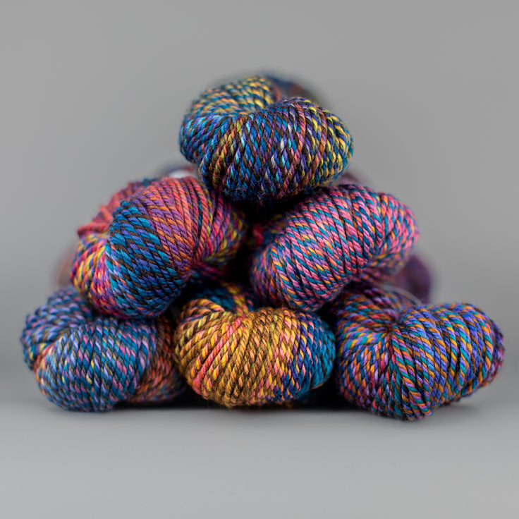 An image of skeins of Spincycle Dream State worsted weight yarn in a blue, pink, purple, and yellow color gradient against a gray background.