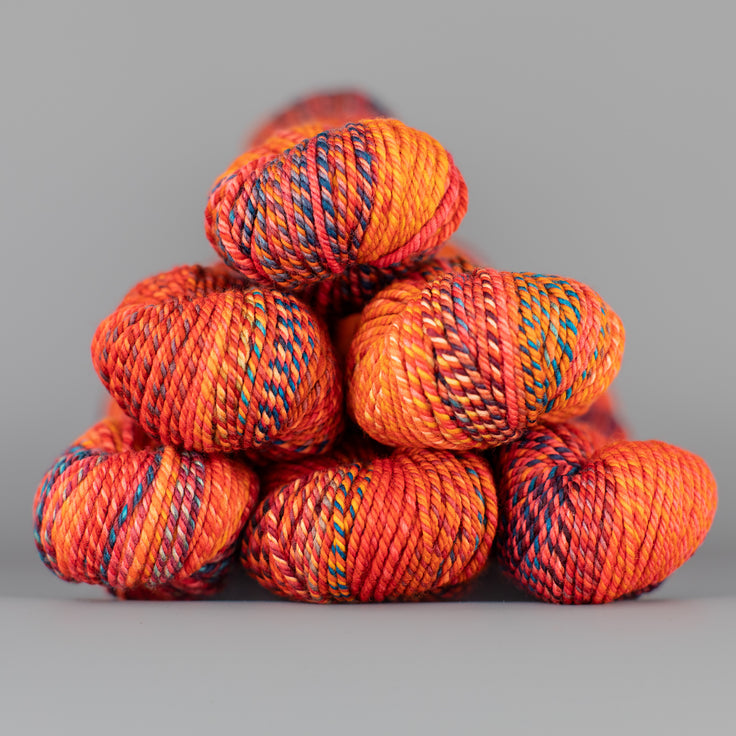An image of skeins of Spincycle Dream State worsted weight yarn in a bright orange, red, and teal color gradient against a gray background.