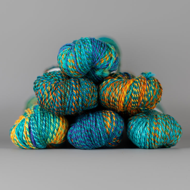 An image of skeins of Spincycle Dream State worsted weight yarn in an aqua blue, orange, and teal color gradient against a gray background.