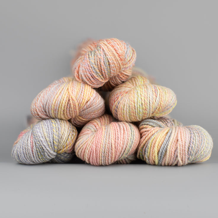 An image of skeins of Spincycle Dream State worsted weight yarn in a pale peach yellow, and taupe color gradient against a gray background.