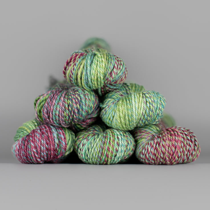 An image of skeins of Spincycle Dream State worsted weight yarn in a sea green, purple pink, and maroon color gradient against a gray background.
