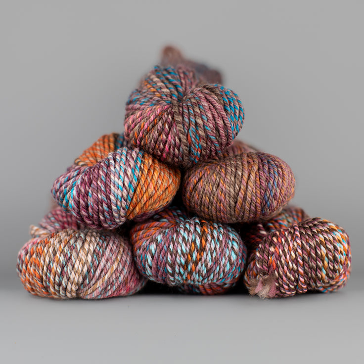 An image of skeins of Spincycle Dream State worsted weight yarn in a blue, brown, mauve, and orange color gradient against a gray background.