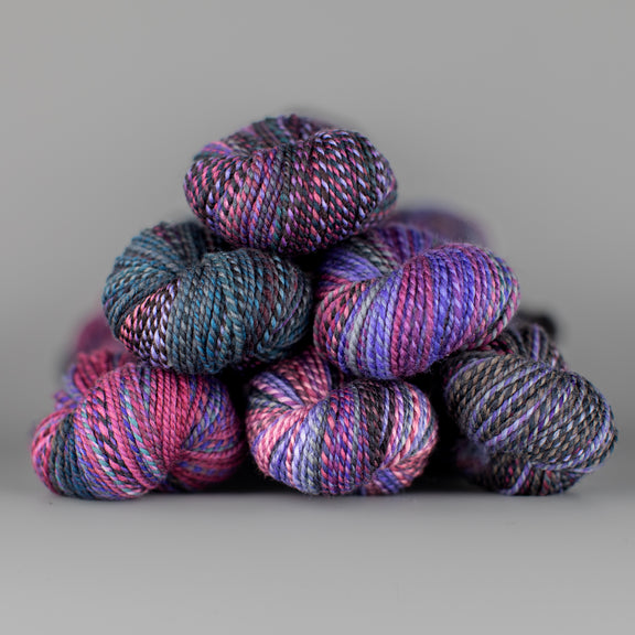 Skeins of Spincycle Dyed in the Wool sport weight yarn in a blue, gray, pink, and purple color gradient against a gray background.