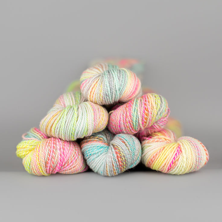 Skeins of Spincycle Dyed in the Wool sport weight yarn in a pale pink, blue, yellow, and gray color gradient against a gray background.