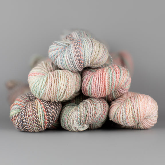 Skeins of Spincycle Dyed in the Wool sport weight yarn in a pale pink, blue, and brown color gradient against a gray background.