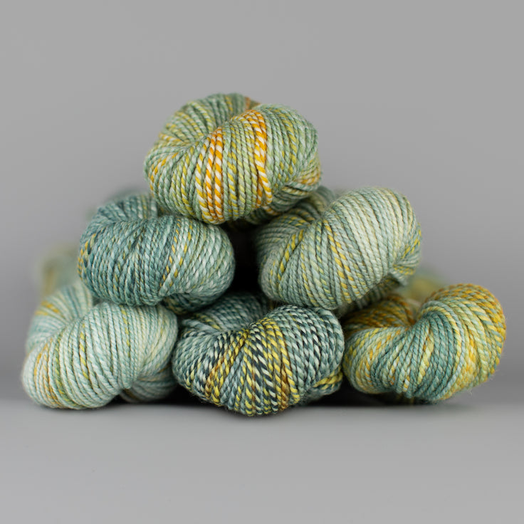 Skeins of Spincycle Dyed in the Wool sport weight yarn in a green, yellow, and aqua blue color gradient against a gray background.