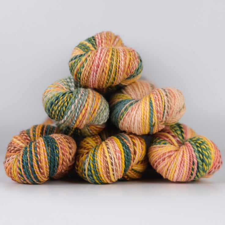 Skeins of Spincycle Dyed in the Wool sport weight yarn in a yellow orange, pink, and green color gradient against a gray background.
