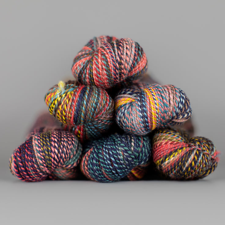 Skeins of Spincycle Dyed in the Wool sport weight yarn in a pink, yellow, blue, brown, and black color gradient against a gray background.