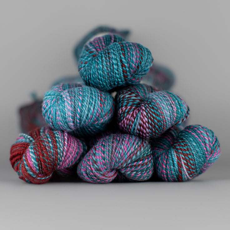 Skeins of Spincycle Dyed in the Wool sport weight yarn in a blue, red, and light purple color gradient against a gray background.
