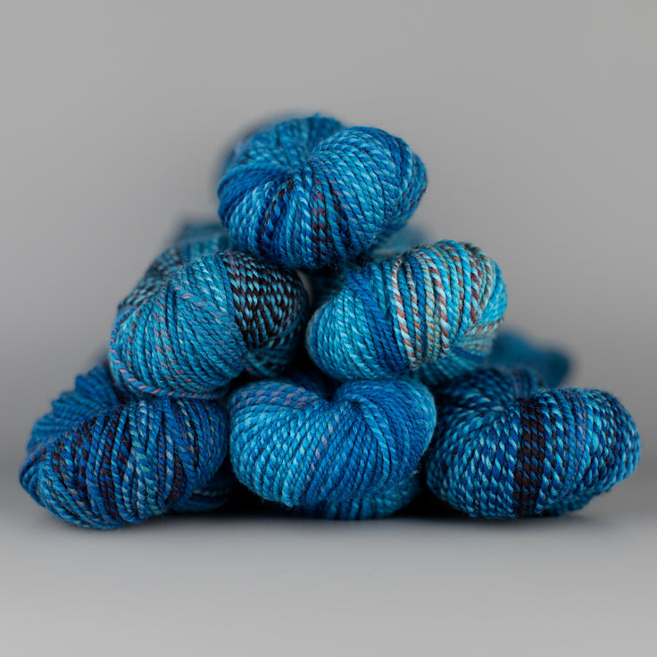 Skeins of Spincycle Dyed in the Wool sport weight yarn in a blue, gray, and deep purple color gradient against a gray background.