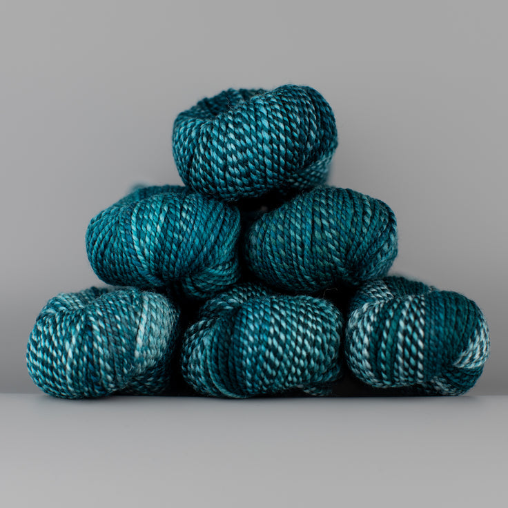 Skeins of Spincycle Dyed in the Wool sport weight yarn in a teal, navy, and gray blue color gradient against a gray background.