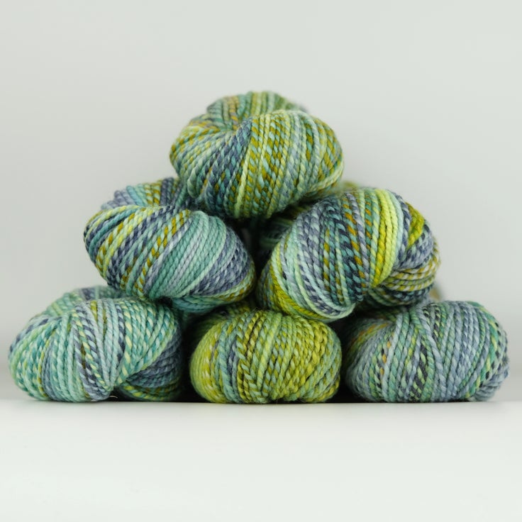 Skeins of Spincycle Dyed in the Wool sport weight yarn in a green, blue, and gray color gradient against a gray background.