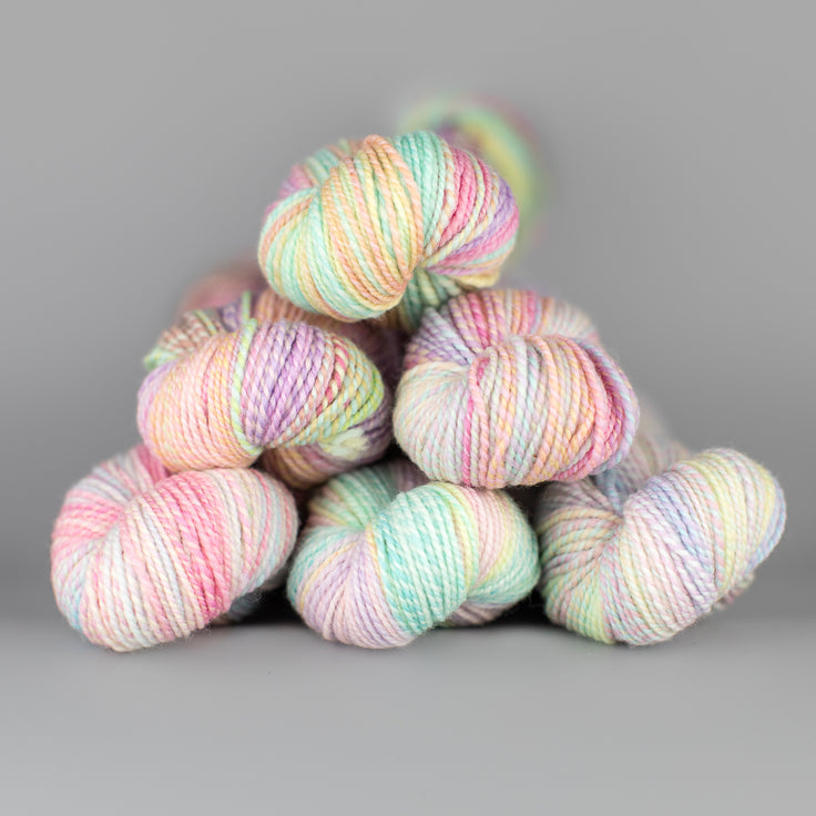 Skeins of Spincycle Dyed in the Wool sport weight yarn in a pastel blue, pink, and yellow color gradient against a gray background.