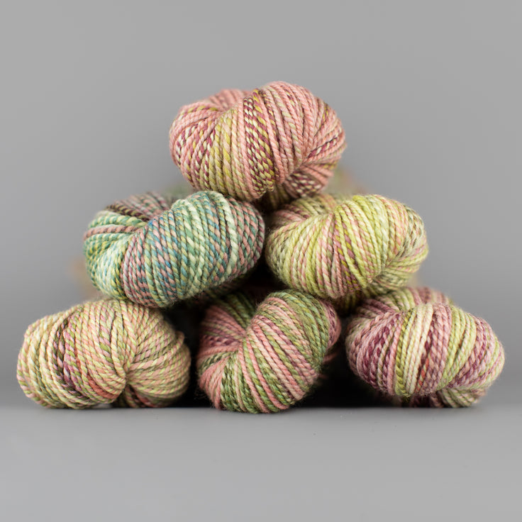 Skeins of Spincycle Dyed in the Wool sport weight yarn in a pale pink, coral, and green color gradient against a gray background.