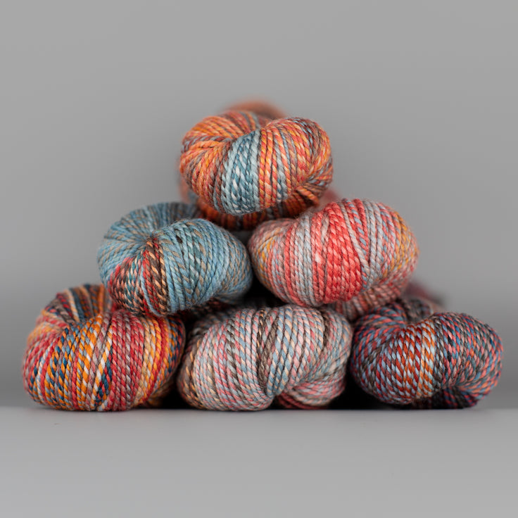 Skeins of Spincycle Dyed in the Wool sport weight yarn in a brown, gray blue, orange,  red, and pink color gradient against a gray background.