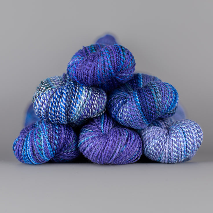 Skeins of Spincycle Dyed in the Wool sport weight yarn in a purple, blue, and gray color gradient against a gray background.