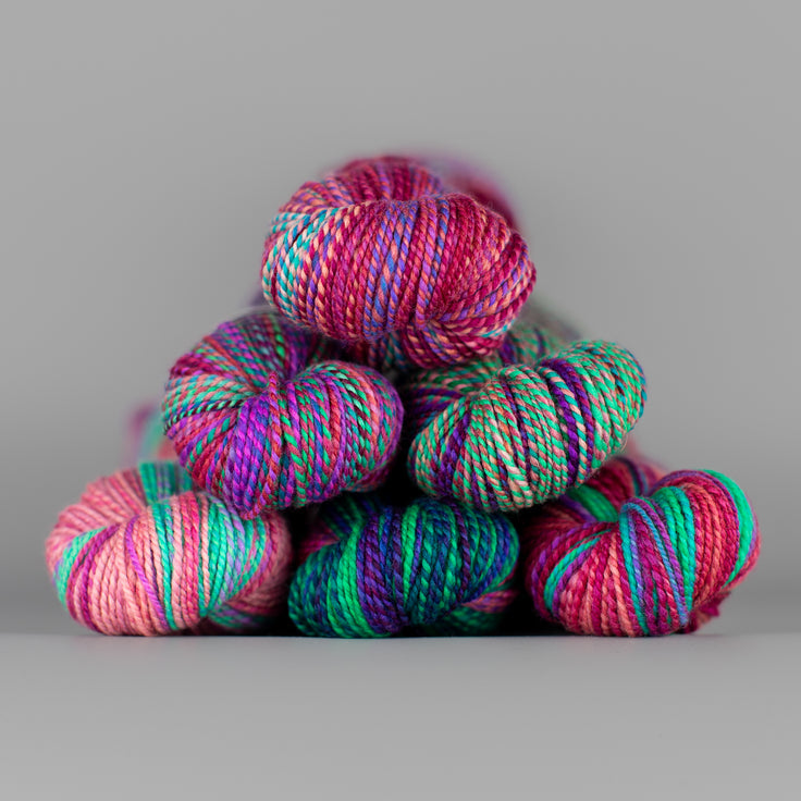Skeins of Spincycle Dyed in the Wool sport weight yarn in a sea green, magenta, teal, and peach color gradient against a gray background.