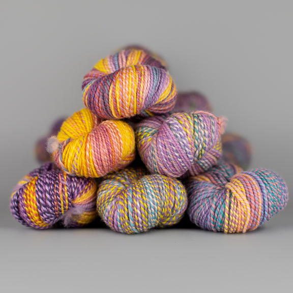 Skeins of Spincycle Dyed in the Wool sport weight yarn in a yellow orange, light purple, and blue color gradient against a gray background.