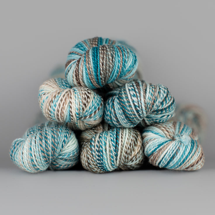 Skeins of Spincycle Dyed in the Wool sport weight yarn in a gray, sky blue, and teal color gradient against a gray background.