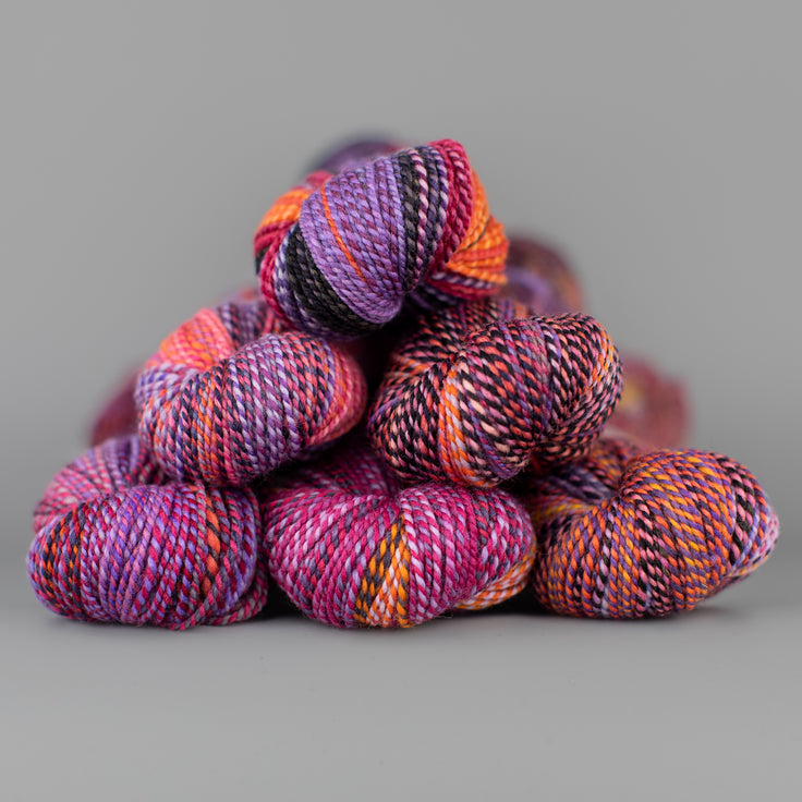Skeins of Spincycle Dyed in the Wool sport weight yarn in an orange, pink, purple, and charcoal color gradient against a gray background.