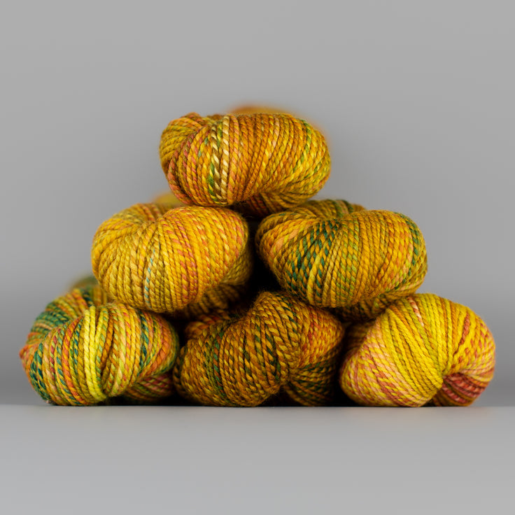 Skeins of Spincycle Dyed in the Wool sport weight yarn in a yellow, brown, and dark green color gradient against a gray background.