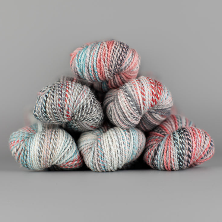 Skeins of Spincycle Dyed in the Wool sport weight yarn in a coral, gray, and pale blue color gradient against a gray background.