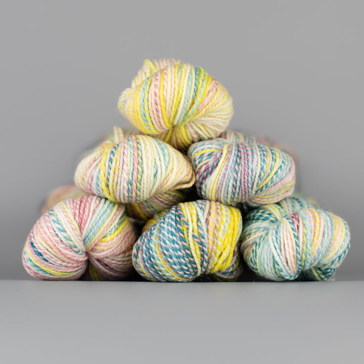 Skeins of Spincycle Dyed in the Wool sport weight yarn in a pale yellow, pale pink, and teal color gradient against a gray background.