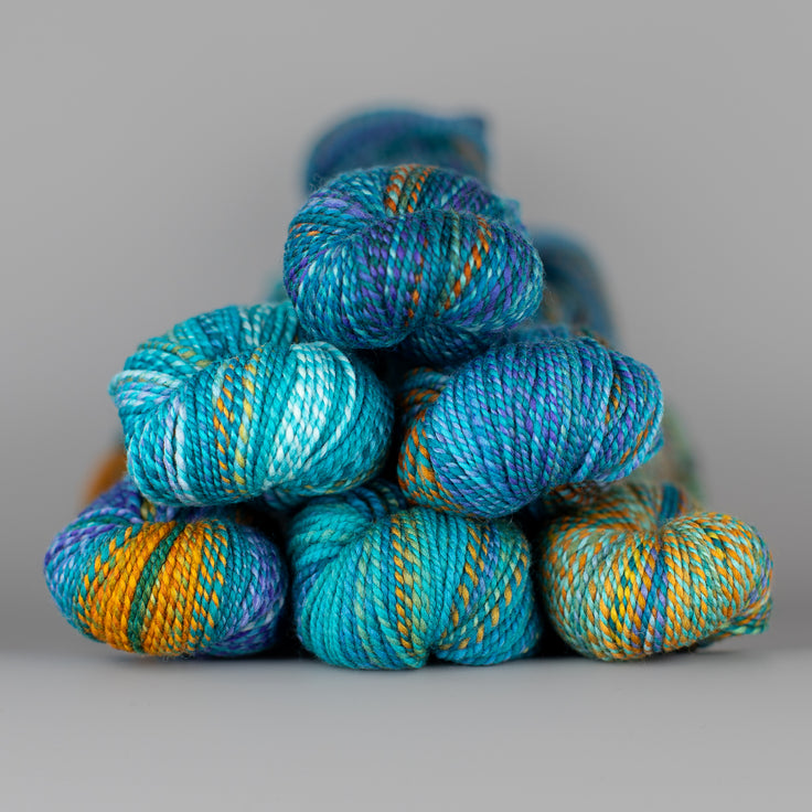 Skeins of Spincycle Dyed in the Wool sport weight yarn in a blue, purple, orange, and green color gradient against a gray background.