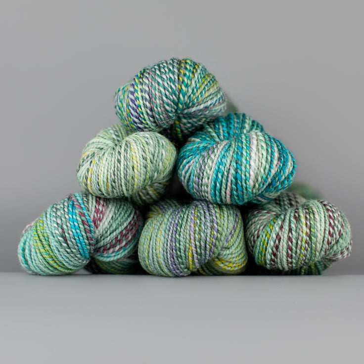 Skeins of Spincycle Dyed in the Wool sport weight yarn in a light green, aqua blue, gray, and maroon color gradient against a gray background.